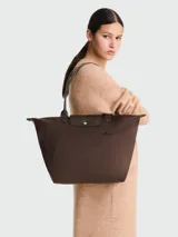 Bolso tote Longchamp modelo Le Pliage Green L, color marrón chocolate, confeccionado en lona de nylon reciclado con detalles en cuero ruso color marrón oscuro. Cuenta con cierre de cremallera y botón a presión, asas cortas para llevar al hombro y es plegable para facilitar su almacenamiento.