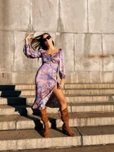 Vestido midi con estampado floral en tonos lila, amarillo y verde. Tiene escote fruncido con lazo ajustable, mangas largas abullonadas y tajo lateral.