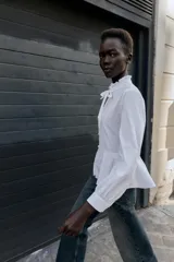 Camisa blanca de popelín con cuello redondo con volante y lazo, manga larga y bajo peplum. Cierre frontal con botones.