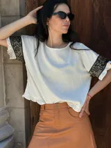 Remera color crudo de corte holgado, con cuello redondo y mangas cortas con volados y apliques de encaje floral negro.