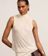 Blusa color crema sin mangas de crepé con cuello alto y lazo por tiras largas de tejido. La prenda tiene cierre por gota con detalle de perla artificial atrás del cuello y frunzidos en la parte delantera.