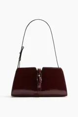 Cartera bandolera color bordó de cuero charolado, con correa fina ajustable y cierre metálico dorado en la parte frontal. Cuenta con un bolsillo interior con cierre y está forrada.