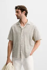 Camisa de hombre relaxed fit confeccionada en tejido de algodón a rayas verticales blancas y negras. Tiene cuello bowling, manga corta, bajo con aberturas laterales y cierre frontal de botonadura.