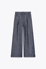 Pantalón ancho azul con textura de lino, tiro alto, cintura elástica con cordón ajustable, pinzas delanteras, bolsillos laterales y traseros, y cinturón de la misma tela con apliques metálicos.