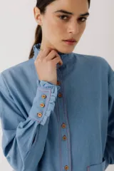 Camisa celeste de jean con cuello alto con volados y mangas largas abullonadas con puños con volados. Presenta botones de madera y costuras rojas a contraste.