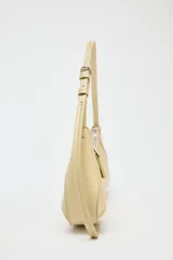 Bolso de hombro color crema, con forma alargada y asa ajustable con hebilla. Tiene pespuntes en el cuerpo, bolsillo interior con cremallera y cierre principal con cremallera y tirador.