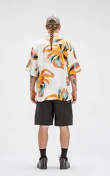 Camisa de manga corta con estampado floral en tonos amarillos, naranjas, azules y marrones sobre fondo blanco. Calce oversize y canesú en la espalda.