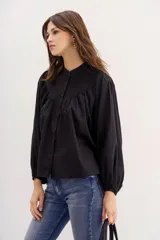 Camisa de corte amplio, color negro, confeccionada en algodón, con cuello redondo, botones al frente y mangas largas abullonadas con puños elastizados.