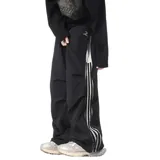 Pantalón deportivo color beige claro, estilo baggy o ancho, con cordón ajustable en la cintura y cierre completo impermeable (full zip) en los laterales. Presenta una franja vertical negra con tres líneas blancas a lo largo de la costura lateral, y un pequeño logo bordado en la parte superior del muslo.