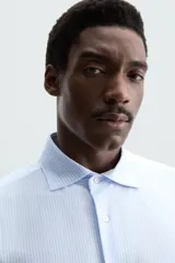Camisa celeste a rayas verticales blancas, de corte recto, con cuello italiano abierto, mangas largas con puños abotonados y cierre frontal con botones.