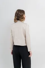 Chaqueta color beige de corte recto, estilo camisa, confeccionada en gamuza con elastano. Presenta cuello camisero, cierre frontal con botones metálicos, bolsillos grandes en el frente y charreteras en los hombros.