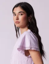 Vestido corto de lino a rayas verticales color lila y blanco, con cuello fruncido, escote en V y lazo de ajuste. Mangas cortas con puntilla de crochet.