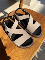 Sandalias bajas color beige con tiras cruzadas y hebilla al tobillo. Suela de goma negra con pespuntes blancos.