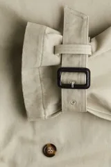 Gabardina midi color beige de algodón, con doble botonadura, cuello con solapas, cierre de corchete en la parte superior, solapa de tormenta en la espalda, bolsillos laterales con solapa, cinturón extraíble con hebilla rectangular, mangas raglán con trabilla ajustable en los puños y abertura trasera.