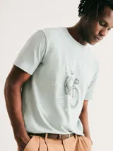 Camiseta blanca de algodón con cuello redondo y mangas cortas. Estampa rectangular con borde negro que contiene la frase "NATURAL ATMOSPHERE" en letras mayúsculas y una imagen de montañas y mar.