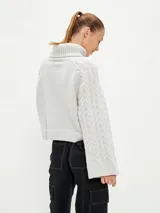 Polerón de tejido de punto color gris claro, con cuello alto y diseño de trenzas en el cuerpo y mangas. Corte holgado y puños anchos.