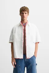 Camisa blanca de algodón con estructura canvas, corte boxy fit, cuello bowling y manga corta. Presenta bordado en azul claro con texto cursivo en el pecho y botones frontales.