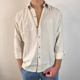 Camisa de manga larga color beige claro, de corte regular, con cuello clásico y cierre frontal con botones. Presenta un pequeño logo bordado en el pecho.