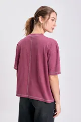 Remera de manga corta color bordó con efecto lavado, corte holgado y costuras externas visibles en blanco en cuello, mangas y ruedo. Presenta un pequeño bordado cursivo en el pecho.