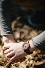 Reloj de pulsera con caja, esfera y malla de madera de sándalo rojo, con marcadores y manecillas negras. Presenta un diseño circular con corona lateral.