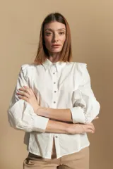 Camisa blanca de manga larga con cuello y botones frontales. Presenta detalles de volados o puntilla en los hombros y mangas abullonadas con puños anchos. La tela parece tener un sutil tejido a rayas verticales.