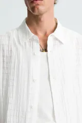 Camisa de hombre de manga corta, cuello solapa y corte relaxed fit. Tejido blanco con textura sutilmente arrugada o plisada. Cierre frontal de botonadura y bajo con aberturas laterales.