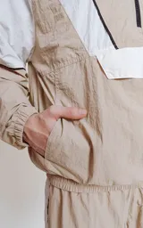 Anorak unisex beige y blanco con capucha, cierre parcial con cremallera, bolsillo tipo canguro con solapa y cordones ajustables con texto en la capucha.