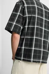 Camisa de hombre relaxed fit con estampado de cuadros blancos sobre fondo negro, cuello solapa, manga corta y cierre frontal con botones.