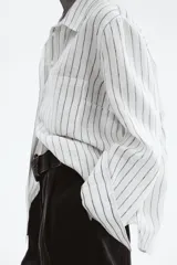 Camisa de lino color blanco con rayas verticales negras, cuello clásico, cierre frontal con botones, manga larga con puños abotonados, bolsillo de parche en el pecho y bajo redondeado.