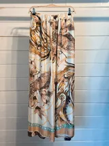 Pantalón ancho de satén con cintura elástica y estampado de motivos florales y cadenas en tonos marrones, beige y dorado sobre fondo blanco. Presenta un borde inferior con detalles en color turquesa.