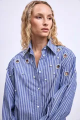 Camisa de mangas largas a rayas verticales azules y blancas, con cuello clásico y cierre frontal con botones. Presenta apliques de strass y ojalillos metálicos.