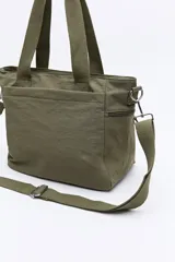 Cartera de nylon color verde militar con asas superiores reforzadas y correa larga regulable y desmontable. Cuenta con bolsillos frontales y laterales con cierre metálico, y un logo de la marca en la parte inferior central.