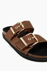 Sandalias planas color tostado con dos tiras anchas sobre el empeine, cada una ajustable con hebillas metálicas doradas.