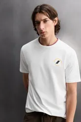 Camiseta blanca de corte holgado confeccionada en tejido de algodón con elasticidad. Tiene cuello redondo, manga corta y estampado en el frente con el logo de la marca y un diseño de tablas de surf.