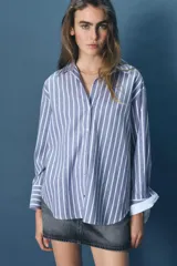 Camisa de algodón a rayas verticales azules y blancas con hilo metalizado, cuello solapa y escote pico. Manga larga acabada en puño, bajo asimétrico y cierre frontal con botones.