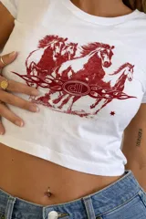 Baby tee blanca de manga corta, corte ajustado, con estampado frontal en color rojo que muestra tres caballos galopando sobre una banda con llamas y un logo central.