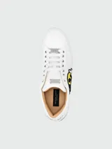 Zapatillas bajas de cuero blanco con aplique de parche bordado de cara sonriente amarilla con relámpagos en los ojos y dedo medio levantado.