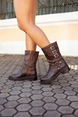 Bota corta de cuero marrón oscuro, estilo motociclista, con caña media arrugada y tres hebillas ajustables en el lateral. Posee una tira decorativa con argolla metálica alrededor del tobillo y suela track gruesa.