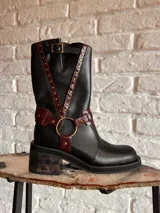 Botas de cuero negro con detalles en color borgoña, hebilla en la parte superior y arnés con aro metálico.