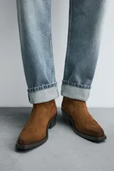 Bota estilo cowboy de cuero color marrón con acabado serraje engrasado, caña media con pespuntes decorativos, tiradores laterales, tacón ancho y punta cuadrada.