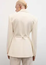Blazer color crema con cuello de solapa, manga larga con puños abotonados, cinturón con hebilla y bolsillos delanteros con solapa.
