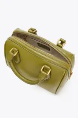 Bolso formato mini bowling color verde oliva con detalles metálicos dorados en las asas. Tiene doble asa de mano y correa bandolera extraíble. Incluye bolsillo interior con cremallera y cierre principal con cremallera.