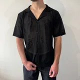 Camisa negra de tejido calado con cuello tipo cubano, manga corta y cierre frontal con botones.