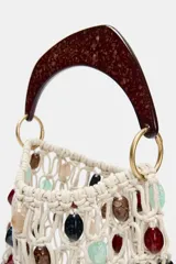 Bolso de mano tipo saca, tejido en red de color crudo, adornado con cuentas ovaladas de colores variados (rojo, negro, verde menta, beige). Posee un asa rígida de mano con acabado carey marrón oscuro.