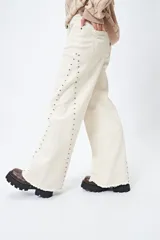 Pantalón de denim blanco con calce wide leg y ruedo desflecado. Presenta tachas metálicas a lo largo de los laterales.