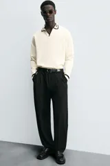Pantalón gris de corte ancho con cintura elástica, pliegues frontales, bolsillos laterales y traseros de vivo. Incluye detalle de hebilla metálica en la trabilla y cierre frontal con cremallera y botón.