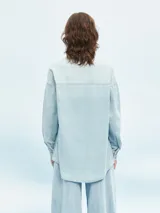 Camisa celeste de denim con botones marrones, corte holgado y mangas largas.