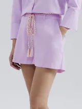 Short de lino color lila claro, de corte recto y tiro medio, con bolsillos laterales. Se complementa con un cinturón trenzado de cuerda en tonos blanco, naranja y rosa pálido con borlas.