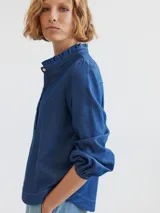 Camisa de denim azul con cuello mao con volados, mangas largas abullonadas con puños elásticos y cierre frontal con botones. Corte semi entallado.