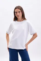 Remera blanca de manga corta con cuello redondo y detalle de ojales metálicos en los puños.